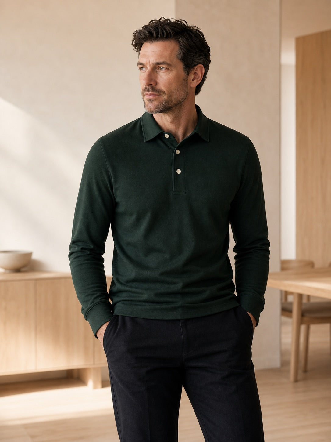 Man in a deep green long-sleeve polo and dark trousers, hands in pockets, in a calm light-wood interior